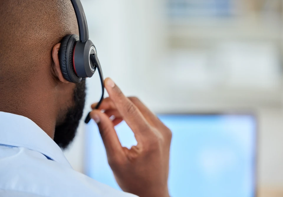 Technicien informatique assis à son bureau, casque audio sur la tête, regardant l'écran de son ordinateur en mode vidéoconférence ou assistance à distance, main sur la souris, environnement de bureau professionnel avec moniteurs et équipements informatiques visibles, concentré et actif en train de fournir du support technique à distance.